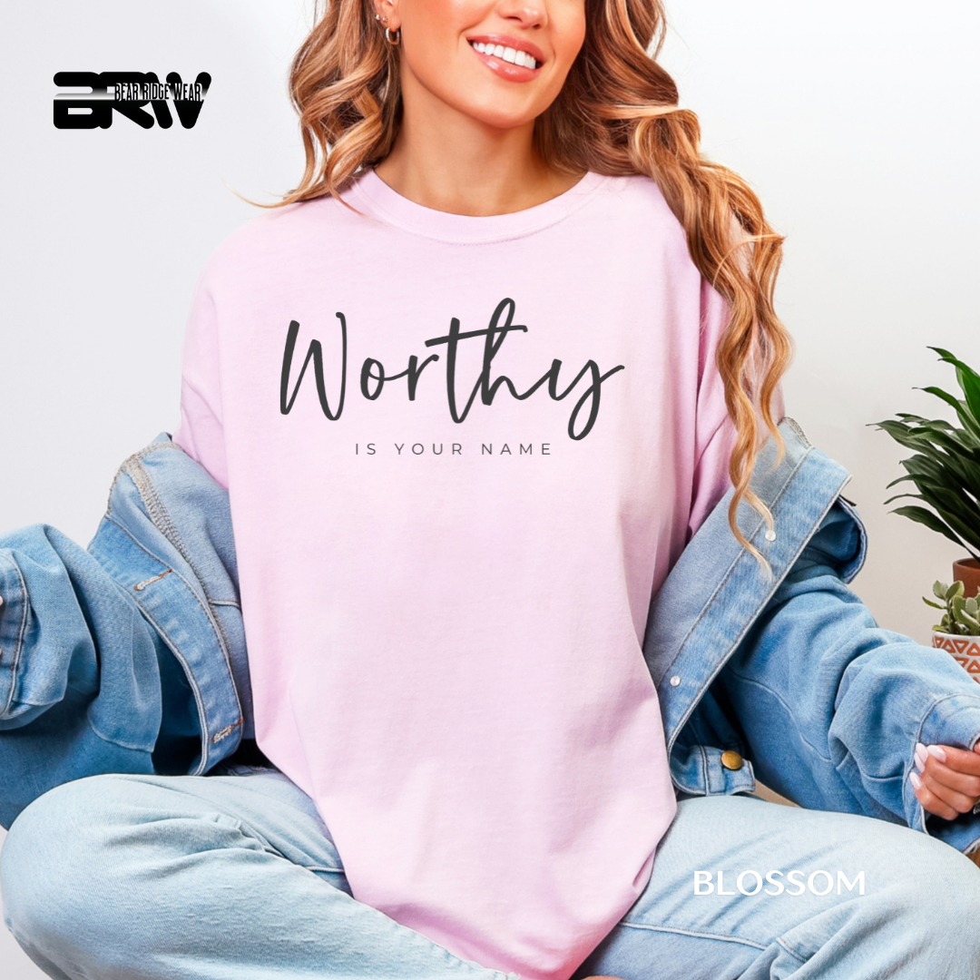 Person wearing a pink sweatshirt with 'Worthy is your name' text, sitting indoors.
