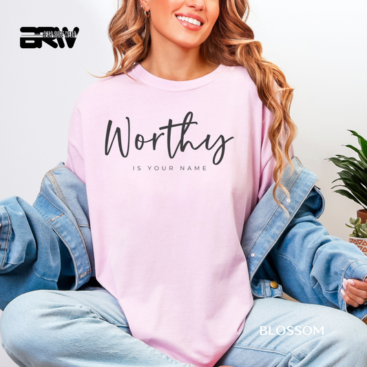 Person wearing a pink sweatshirt with 'Worthy is your name' text, sitting indoors.