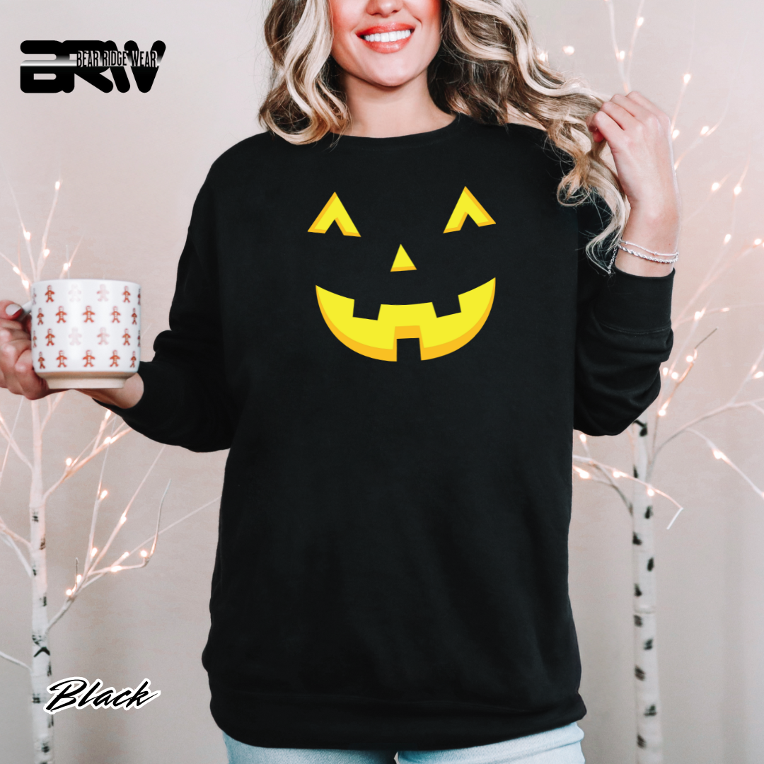 Person wearing a black sweatshirt with a yellow jack-o'-lantern face design, holding a mug with a white background.