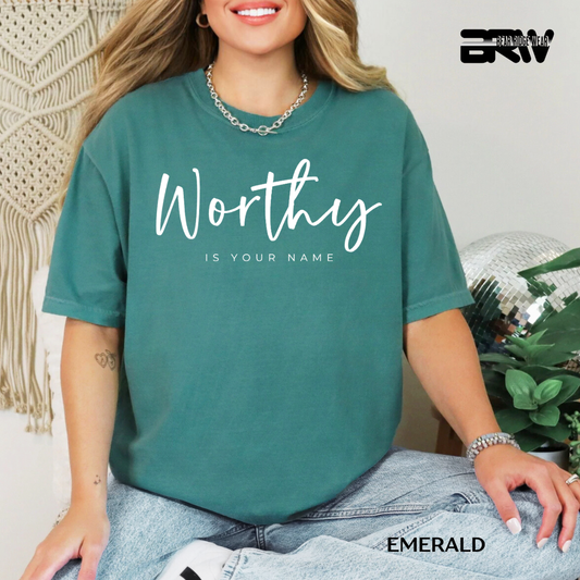 Person wearing a teal t-shirt with 'Worthy is your name' text, sitting indoors.