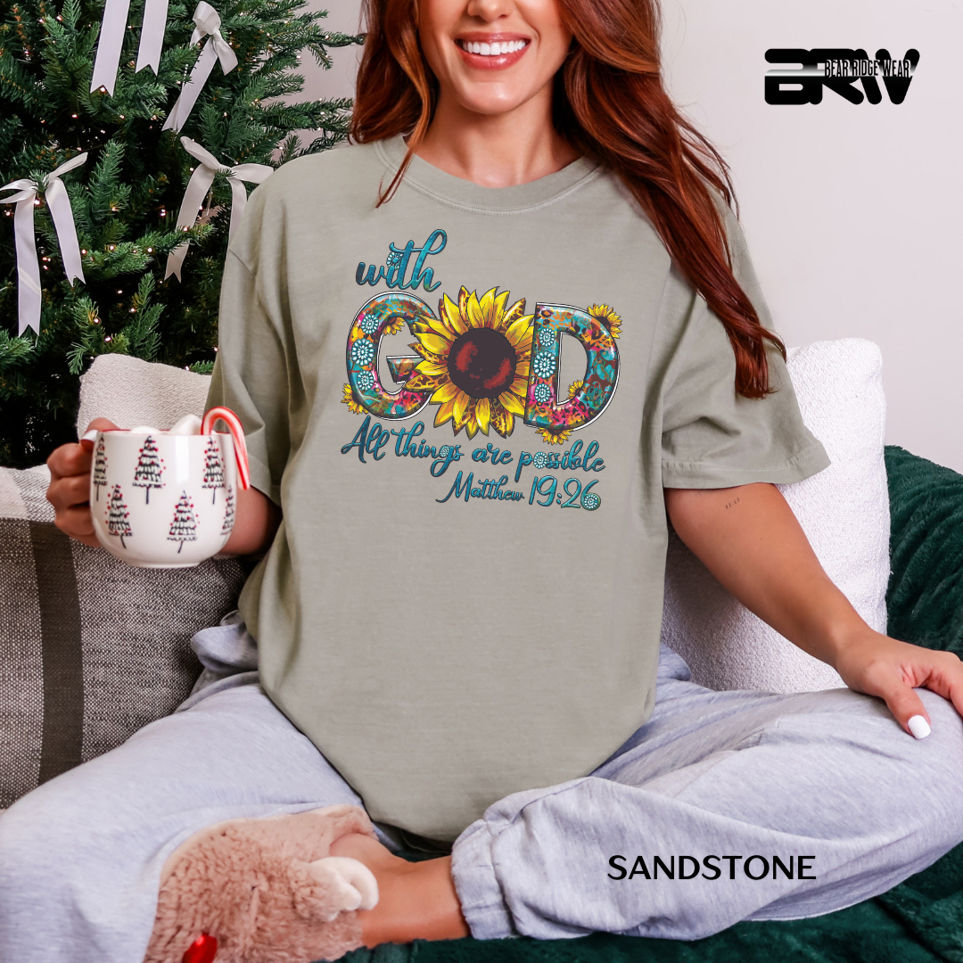 Woman wearing a sandstone t-shirt with a sunflower design and text, holding a mug, sitting on a couch with a Christmas tree in the background.