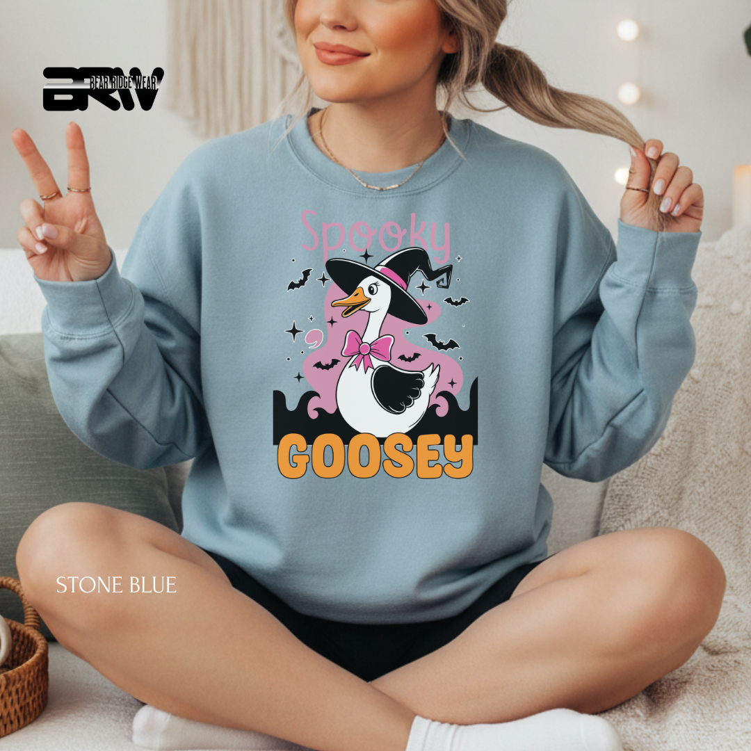Person wearing a stone blue sweatshirt with a goose graphic and text, sitting on a couch.
