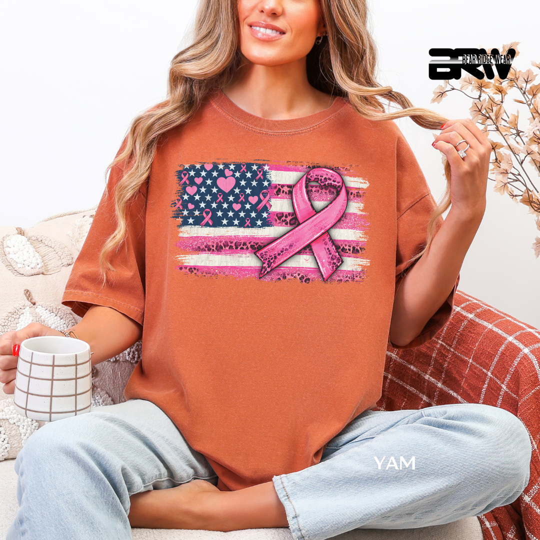 Woman wearing a brown t-shirt with a pink ribbon and American flag design, sitting on a couch.