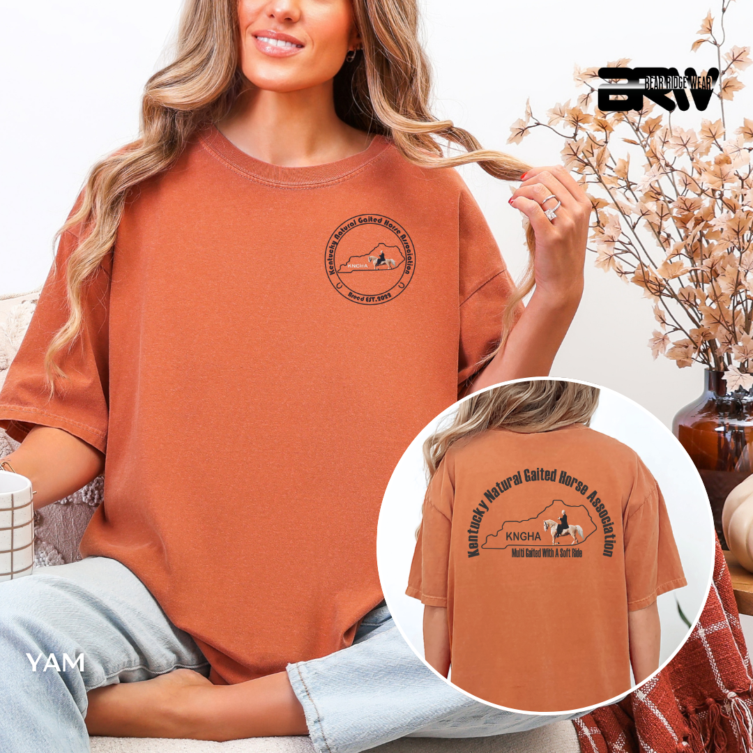 Woman wearing a brown t-shirt with a logo and text, sitting in a cozy room.