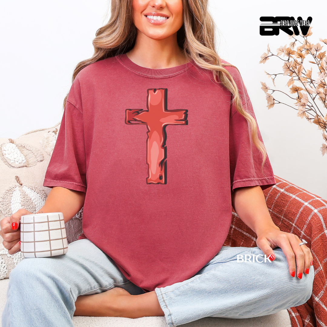 Person wearing a maroon t-shirt with a cross design, sitting on a couch holding a mug.