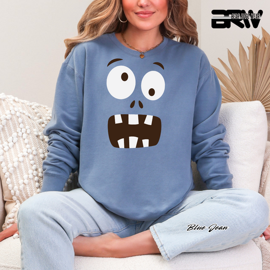 Person wearing a blue sweatshirt with a cartoon face design, sitting on a couch. MONSTER FACE FALL TEE