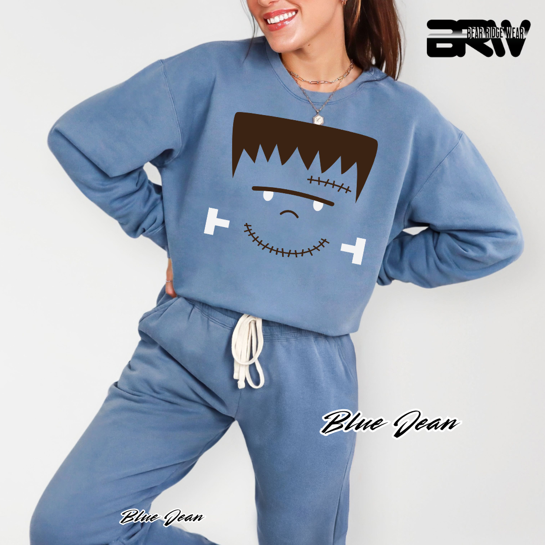 Person wearing a blue sweatshirt with a cartoon face design on a white background, FRANKENTEIN FALL TEE