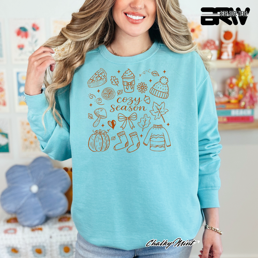 Woman wearing a blue sweater with 'cozy season' design, standing in a colorful room.