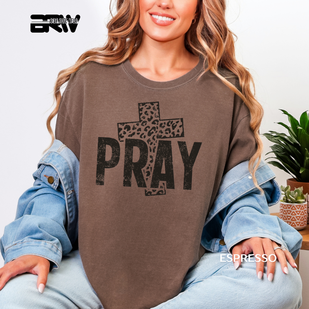 Woman wearing a brown sweatshirt with 'PRAY' and a cross design, sitting indoors.