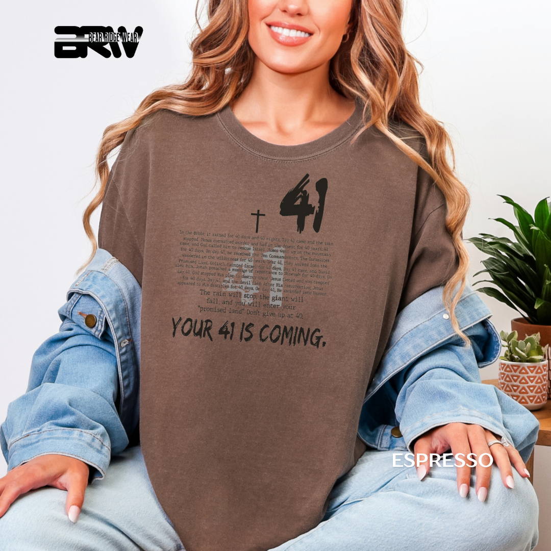 Person wearing a brown sweatshirt with text and a cross design, sitting indoors.