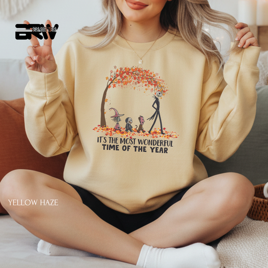 Person wearing a beige sweatshirt with a graphic design and text, sitting on a couch. ITS THE MOST WONDERFUL TIME OF THE YEAR FALL SWEATSHIRT