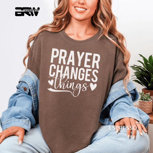 Woman wearing a brown sweatshirt with 'PRAYER CHANGES THINGS' text, sitting indoors.