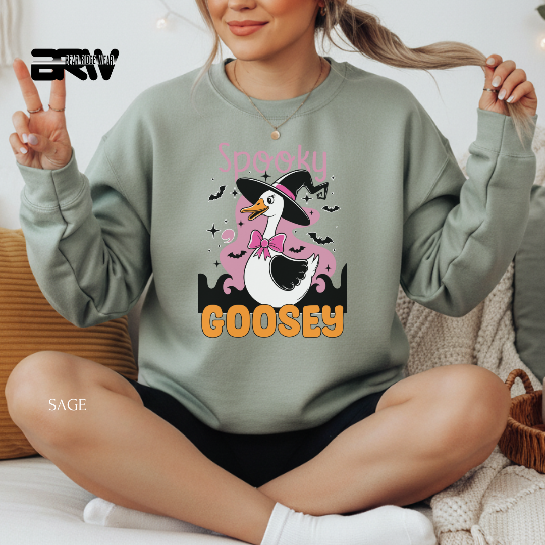 Person wearing a green sweatshirt with a cartoon goose design and text, sitting on a couch. SPOOKY GOOSEY FALL TEE