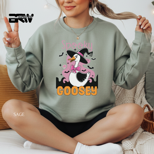 Person wearing a green sweatshirt with a cartoon goose design and text, sitting on a couch. SPOOKY GOOSEY FALL TEE