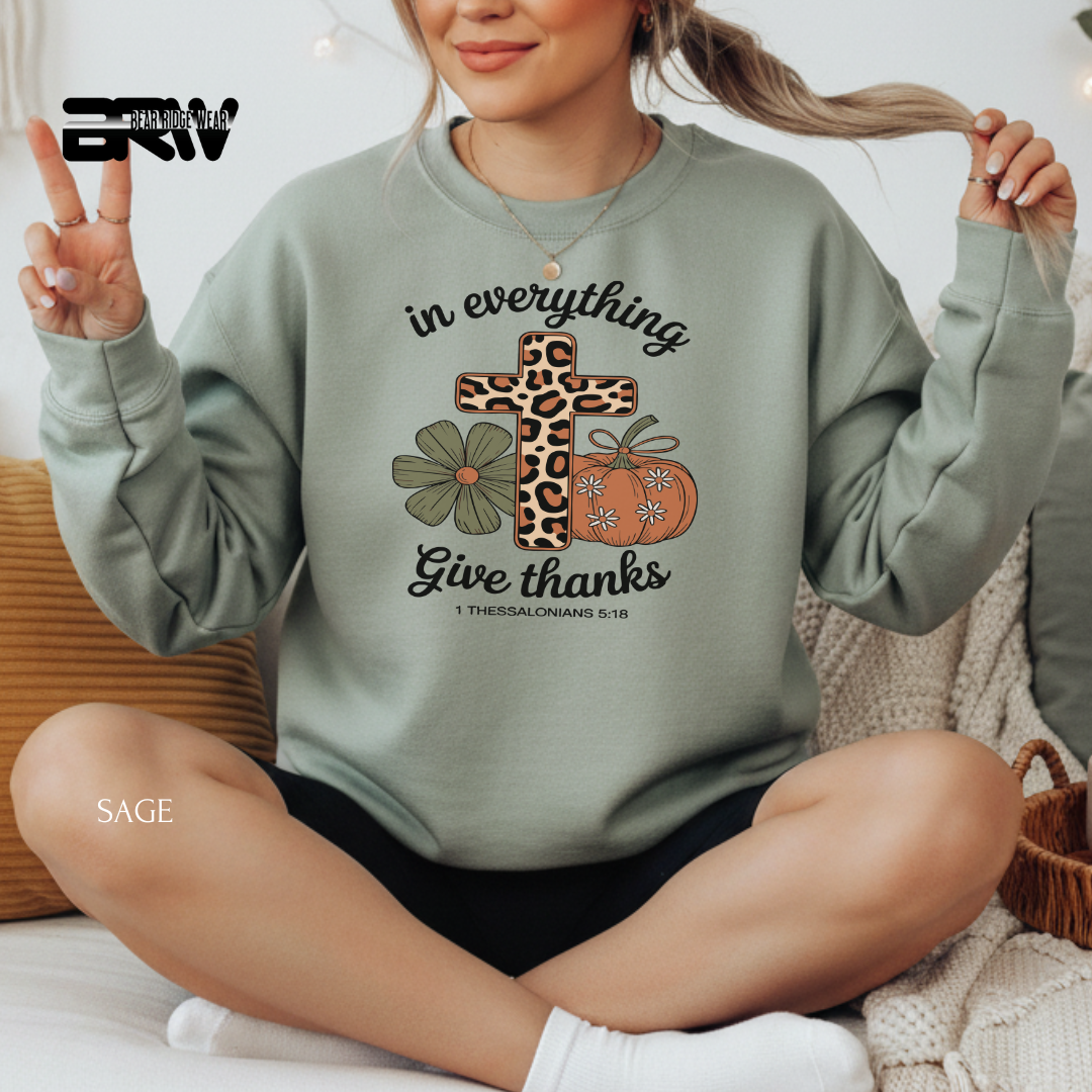 Person wearing a green sweatshirt with a cross, flowers, and pumpkin design, sitting on a couch. IN EVRYTHING GIVE THANKS FALL SWEATSHIRT