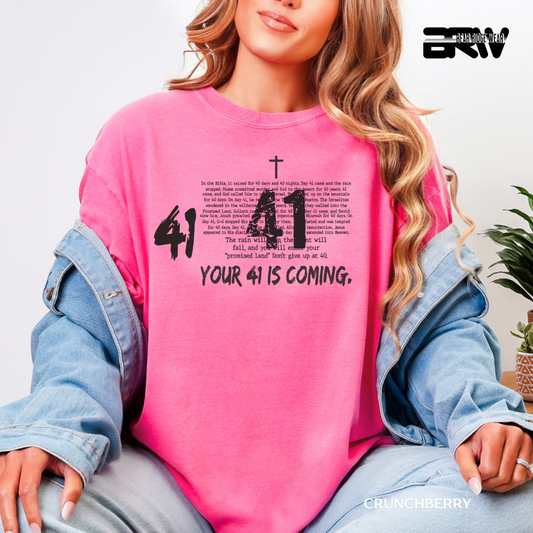 Person wearing a pink sweatshirt with text and a cross design, sitting against a light background.