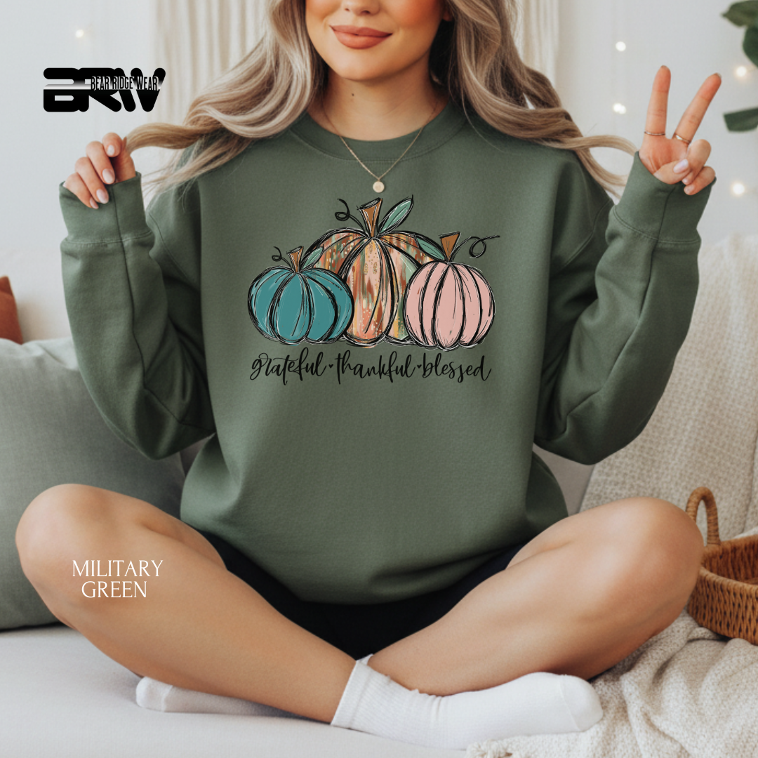 Person wearing a green sweatshirt with pumpkins and text, sitting on a couch. GRATEFUL, THANKFUL, BLESSED FALL SWEATSHIRT