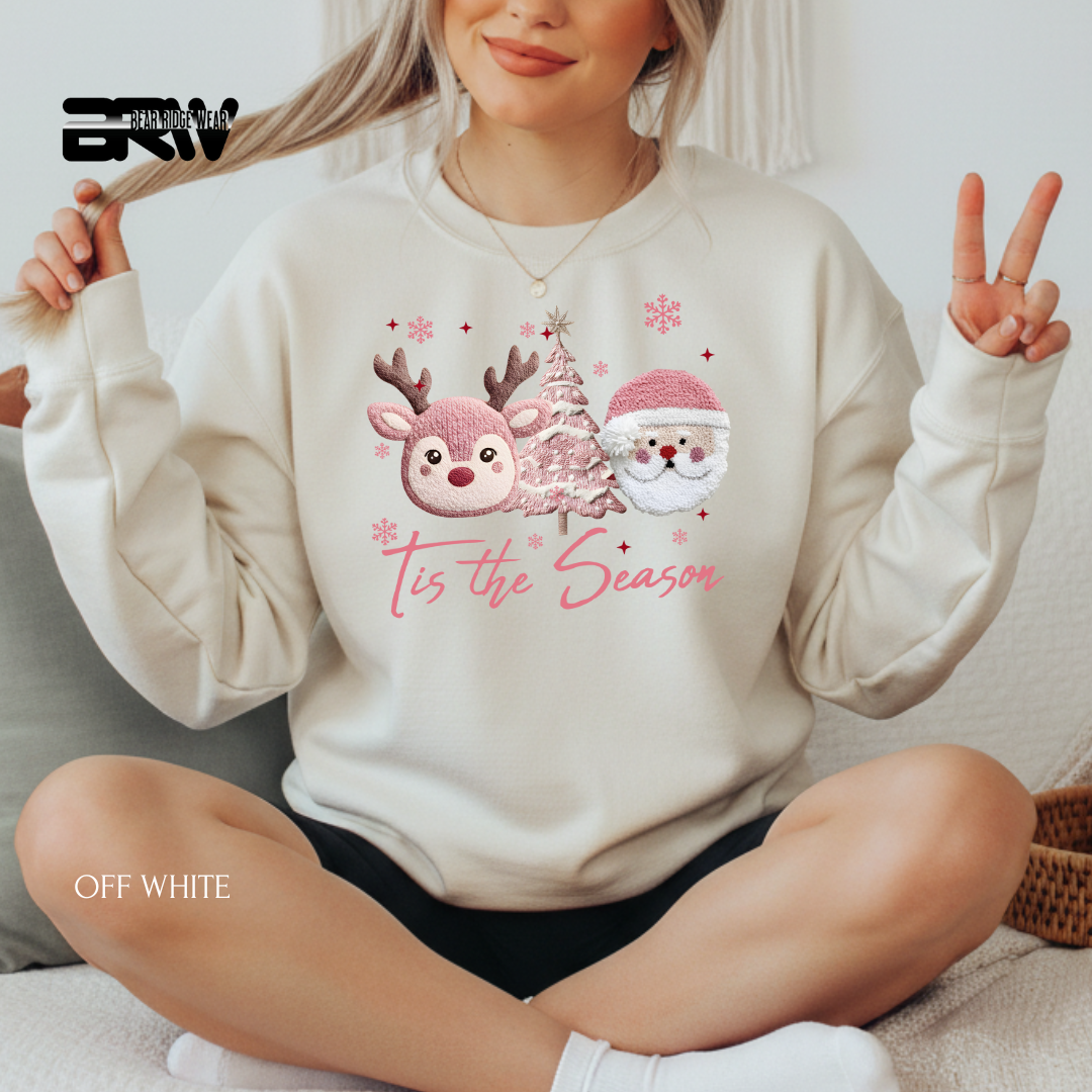 Person wearing a white sweatshirt with Christmas-themed design and text 'Tis the Season', sitting on a couch.