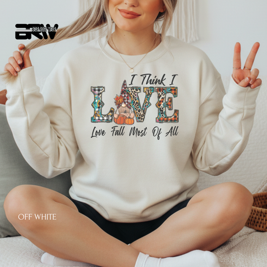 Person wearing a white sweatshirt with colorful text design, sitting on a couch. I THINK I LOVE LOVE FALL MOS OF ALL FALL SWEATSHIRT