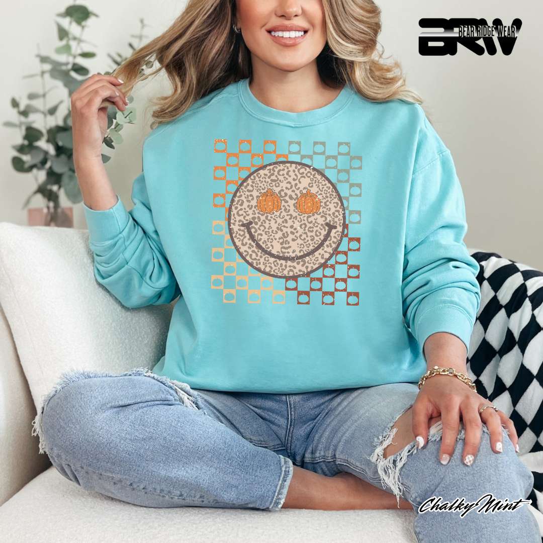 Woman wearing a light blue sweatshirt with a smiley face design, sitting on a couch. FALL SWEATSHIRT 