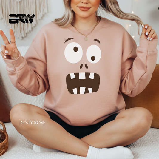 Person wearing a pink sweatshirt with a cartoon face design, sitting on a couch. MONSTER FACE FALL SWEATSHIRT